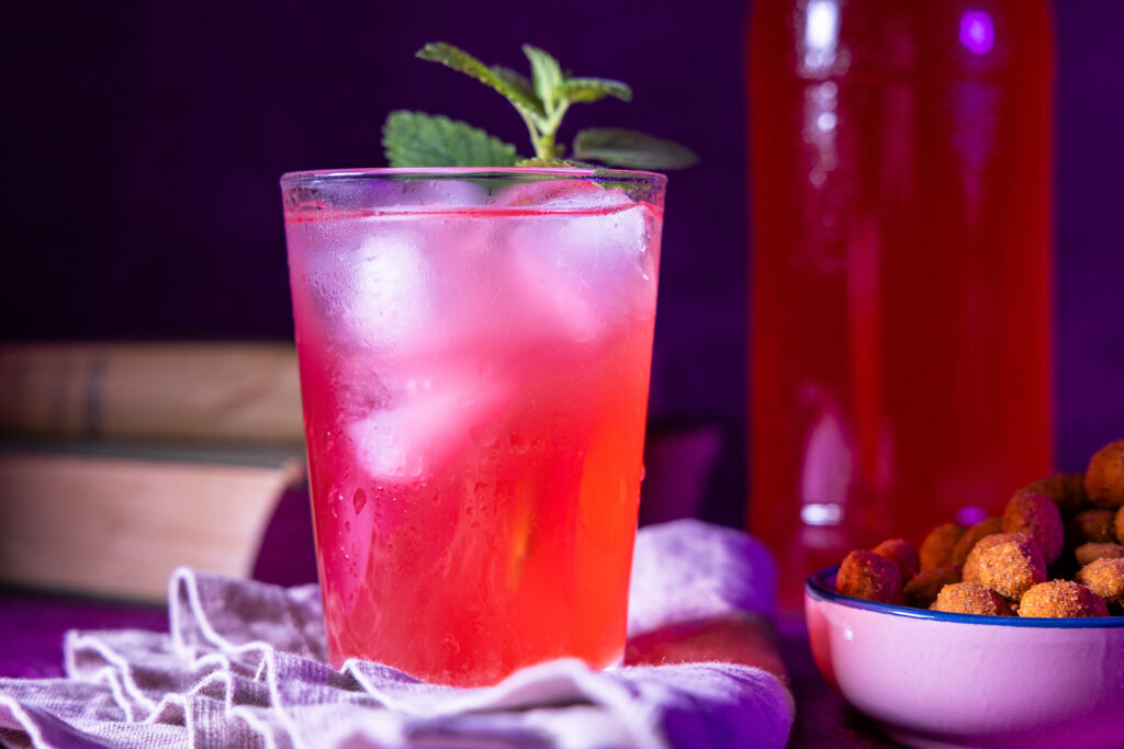Himbeer-Mango-Kefir - selbst fermentiert mit Wasserkefirkristallen. Im Glas mit Eiswürfeln und einem Stengel Melisse. Im Hintergrund eine Flasche Kefir.