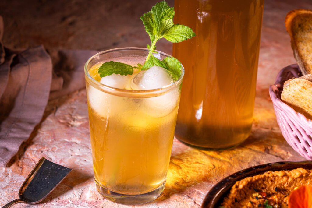 Kombucha-Grundrezept - eine Glas mit Kombucha, einem Stengel Melisse und Eiswürfeln. Dahinter eine Flasche mit Kombucha.