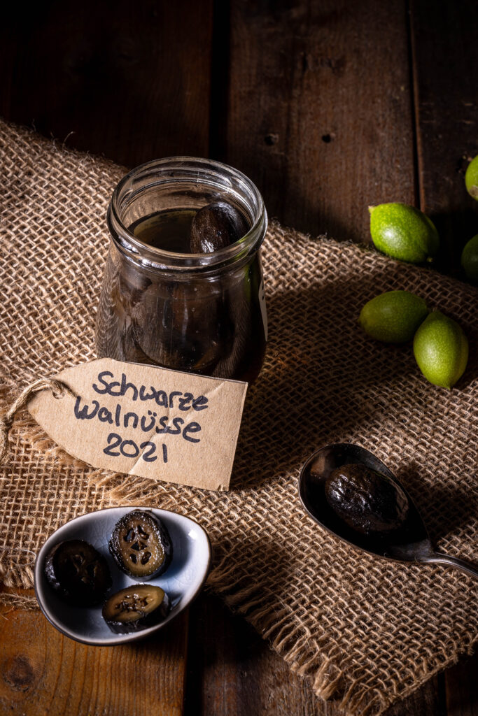 Schwarze Nüsse - im Glas, in einer Schale und auf einem Löffel. Dahinter frische, unreife, grüne Walnüsse.