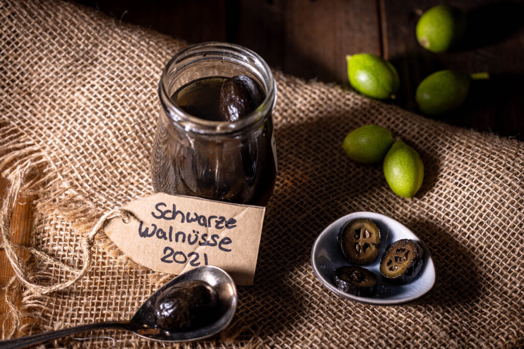 Schwarze Nüsse - im Glas, in einer Schale und auf einem Löffel. Dahinter frische, unreife, grüne Walnüsse.
