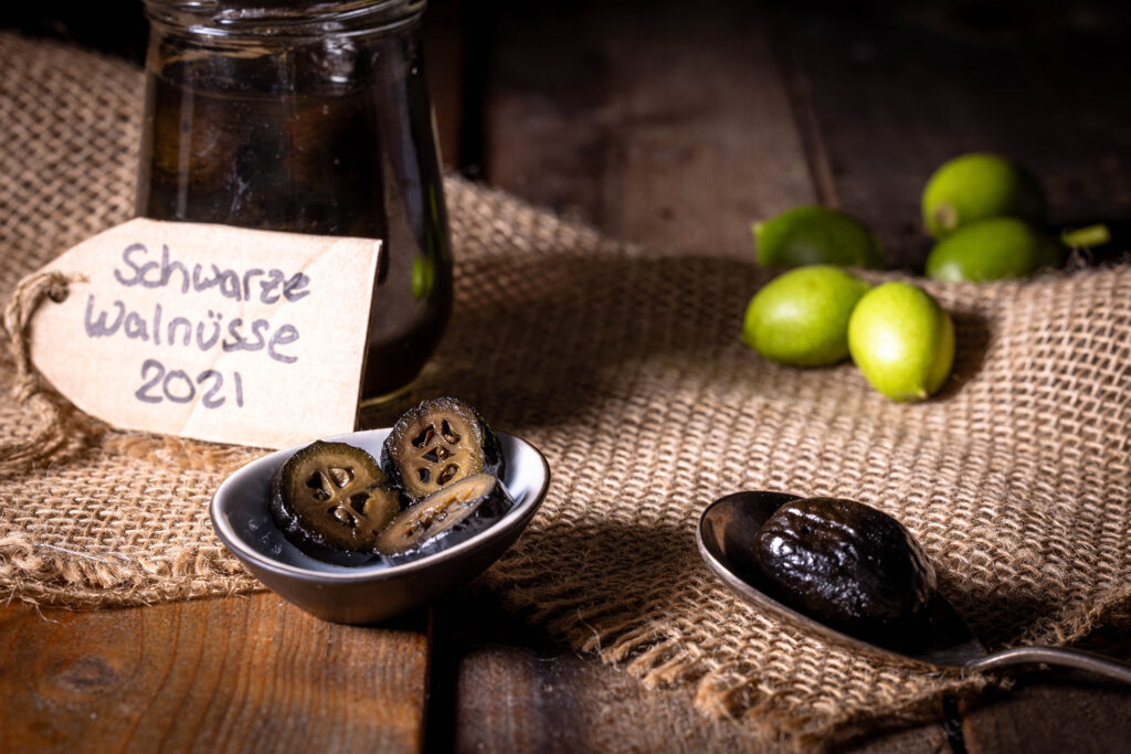 Schwarze Nüsse in Scheiben in einer kleinen Schale und dahinter eingelegt in einem Glas. Vor dem Glas ein Papierschild mit der Aufschrift 'Schwarze Walnüsse 2021'.