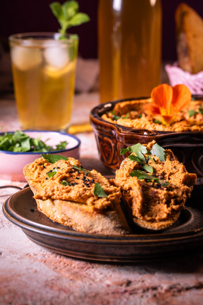 Zwei Brote und eine Schale mit Hummus mit getrockneten Tomaten. Dahinter Kräuter und Getränke.