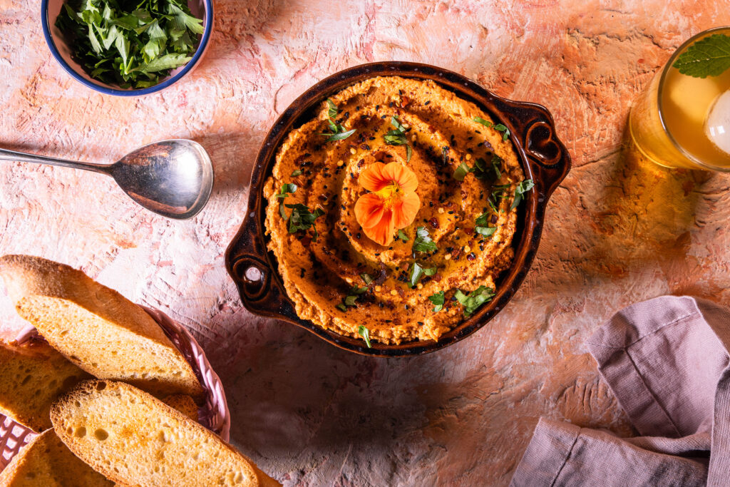 Tomaten-Hummus mit geröstetem Brot Getränk und frischen Kräutern auf einem rötlichen Tisch.