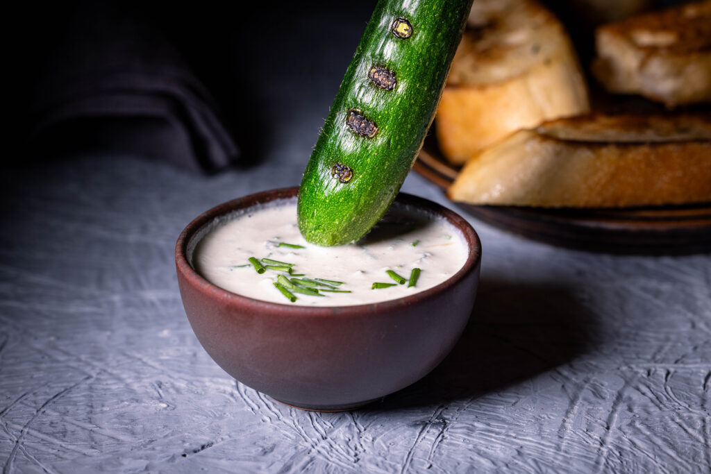 Eine gegrillte Babyzucchini wird in veganen Blauschimmelkäsedip getunkt.