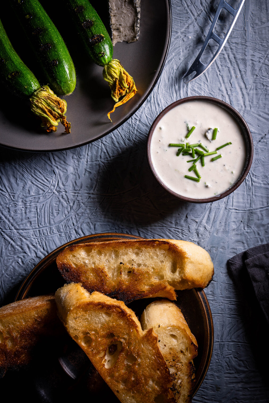 Veganer Blauschimmelkäsedip– in einer Schale, daneben ein Teller mit gerösteten Babyzucchini und ein Teller mit geröstetem Brot.