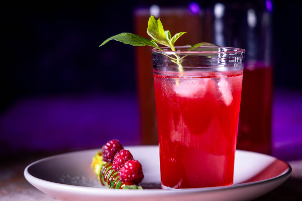 Himbeer-Kardamom-Kombucha in einem Glas mit Eiswürfeln. Daneben frische Himbeeren.