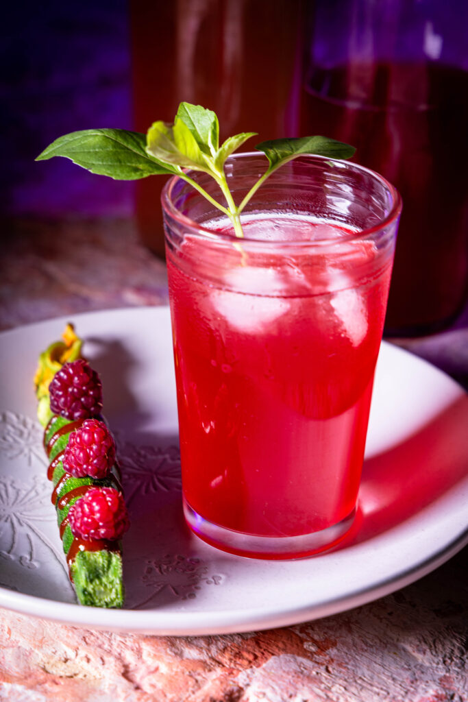 Himbeer-Kardamom-Kombucha in einem Glas mit Eiswürfeln. Daneben Kombucha in Flaschen. Davor frische Himbeeren auf gerösteter Zucchini.