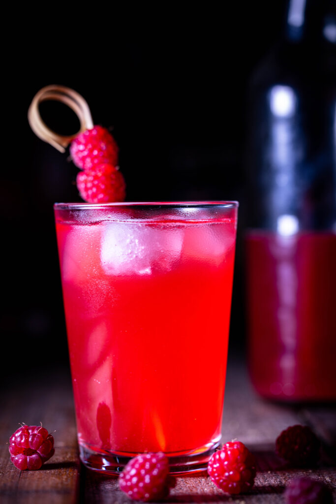 HImbeerkefir mit Eiswürfeln im Glas. Um das Glas auf dem Holztisch: frische Himbeeren. Dahinter eine angebrochene Flasche HImbeerkefir.