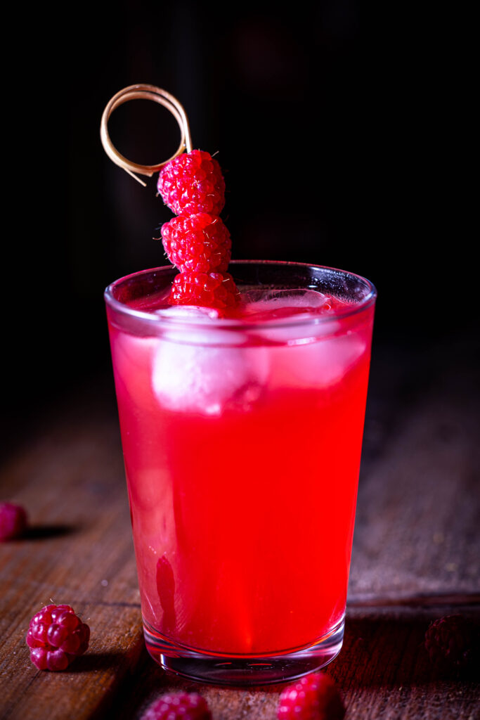 HImbeerkefir mit Eiswürfeln im Glas. Um das Glas auf dem Holztisch: frische Himbeeren. Dahinter eine angebrochene Flasche HImbeerkefir.