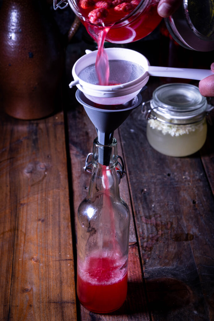 Himberkefir aus Wasserkefir-Zweitfermentation wird durch ein Sieb in eine Flasche abgeseiht.