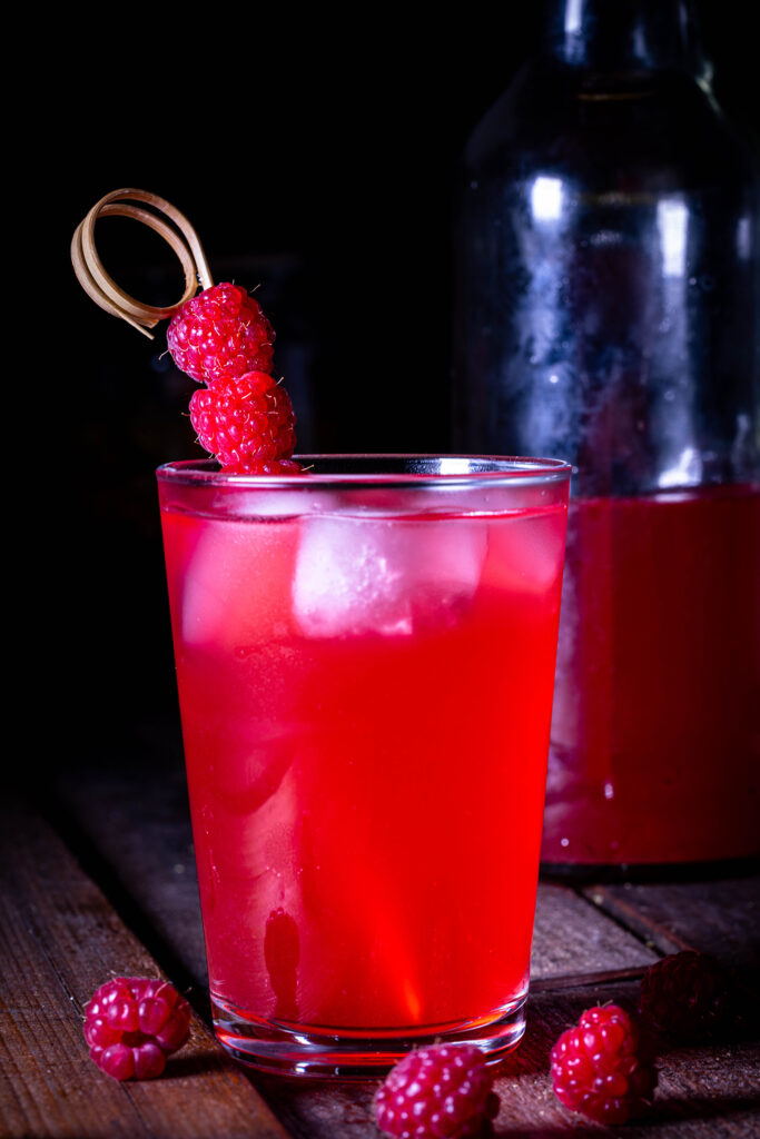 HImbeerkefir mit Eiswürfeln im Glas. Um das Glas auf dem Holztisch: frische Himbeeren. Dahinter eine angebrochene Flasche HImbeerkefir.