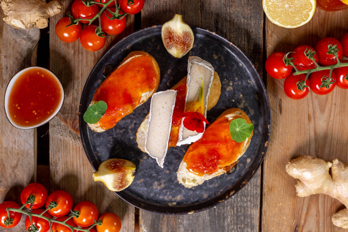 Tomatenkonfitüre auf Brotscheiben, teilweise mit veganem Camembert. Drumrum Cherrytomaten und eine Schale mit Tomatenkonfitüre.