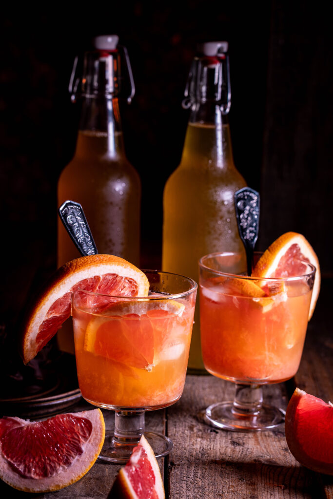 Grapefruit-Kombucha -in Gläsern mit Eis und frischer Grapefruit. Dahinter Kombucha in Flaschen.