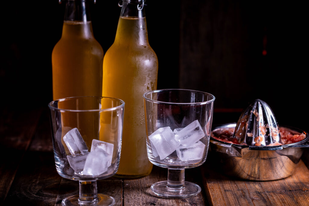 2 Flaschen Kombucha, 2 Gläser mit Eiswürfeln, eine Saftpresse