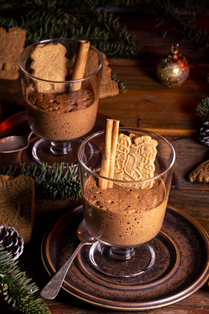 Spekulatius-Mousse in Dessertgläsern, dekoriert mit Zimtstange und einem Spekulatiuskeks. Auf einem weihnachtlichen Holztisch.