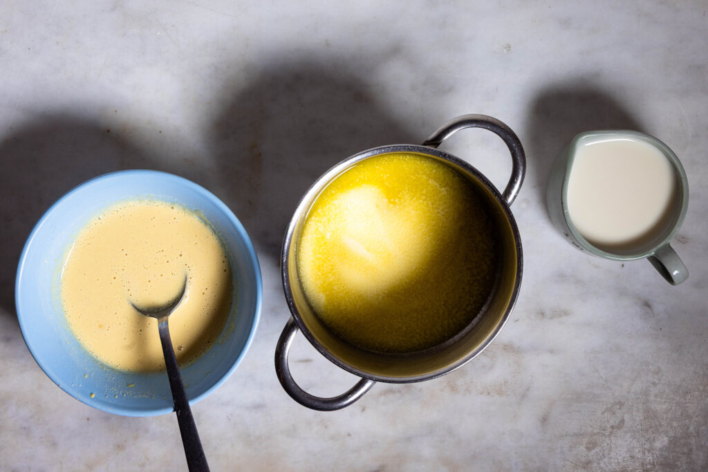 Geschmolzene, vegane Butter in einem kleinen Topf. Daneben Schalen mit Sojamilch und angerührtem Kichererbsenmehl.