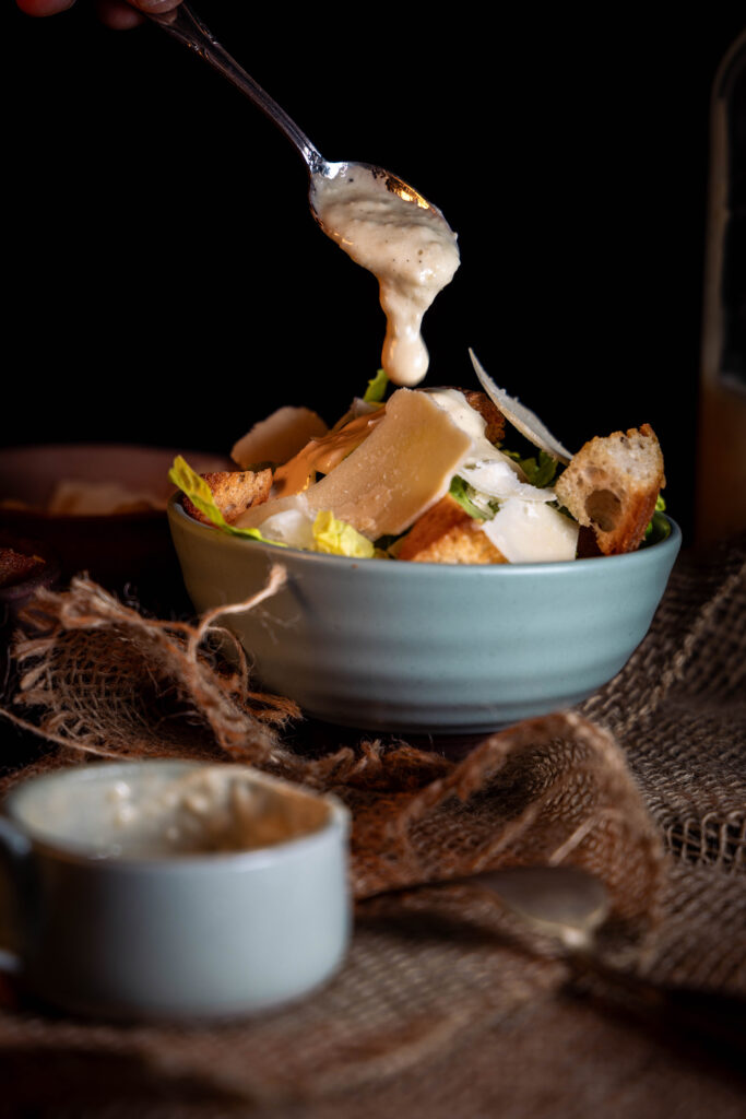 Veganes Caesar Dressingwird mit einem Löffel auf einen Salat gegeben.