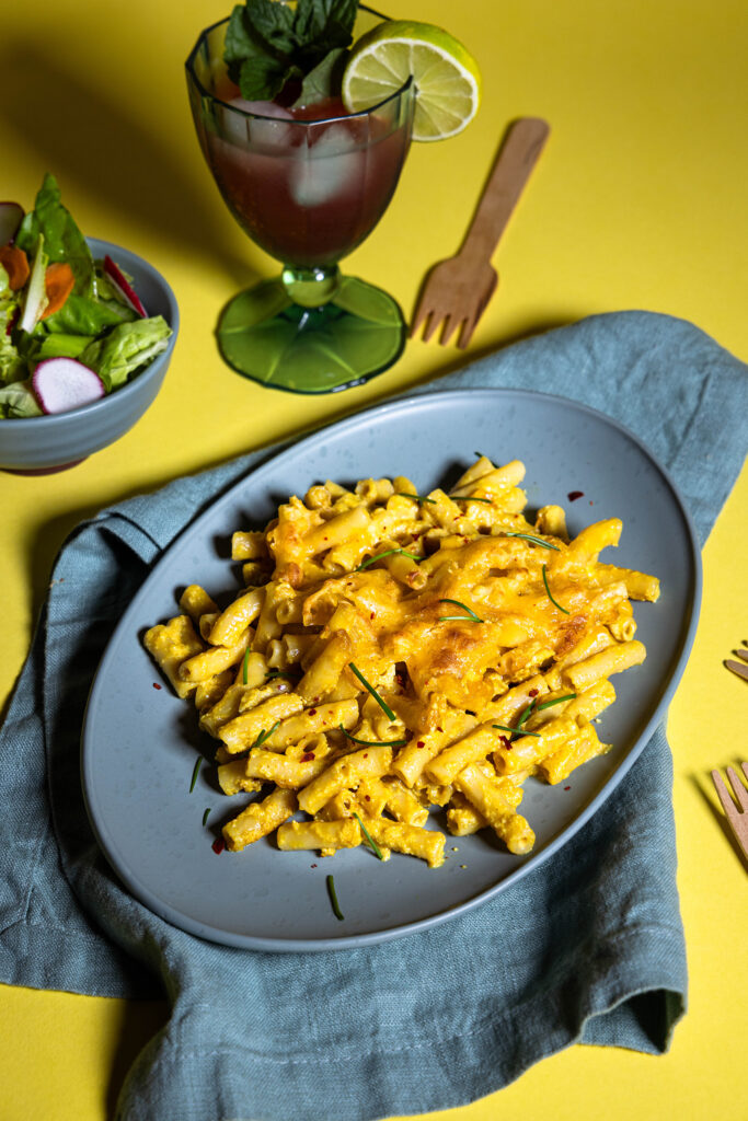 Vegane Maccaroni and Cheese auf einem Teller, außerdem ein Getränk und Salat