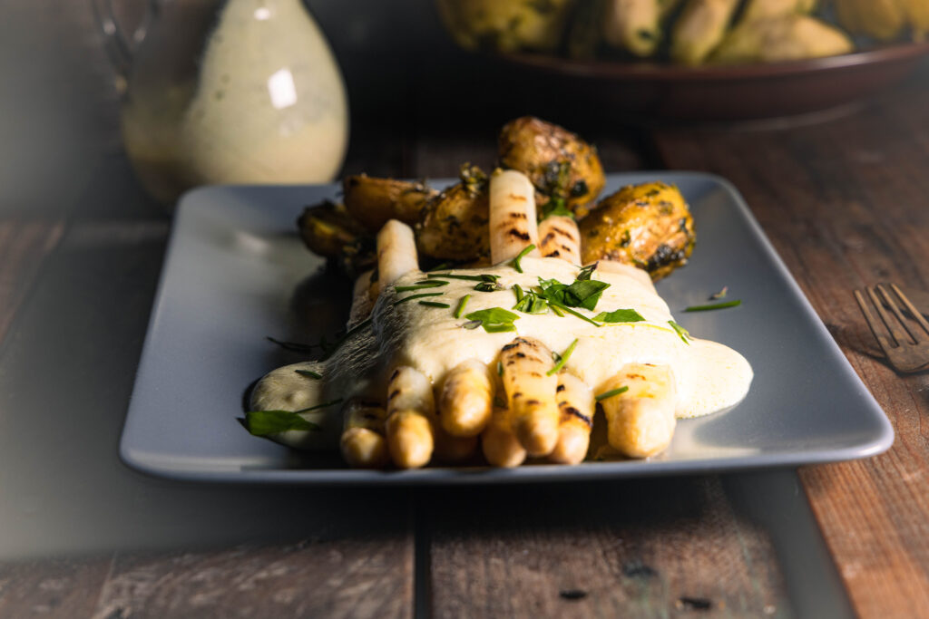 Gegrillter, weißer Spargel mit veganer Sauce Hollandaise, Kräutern und Ofenkartoffeln auf einem Teller.