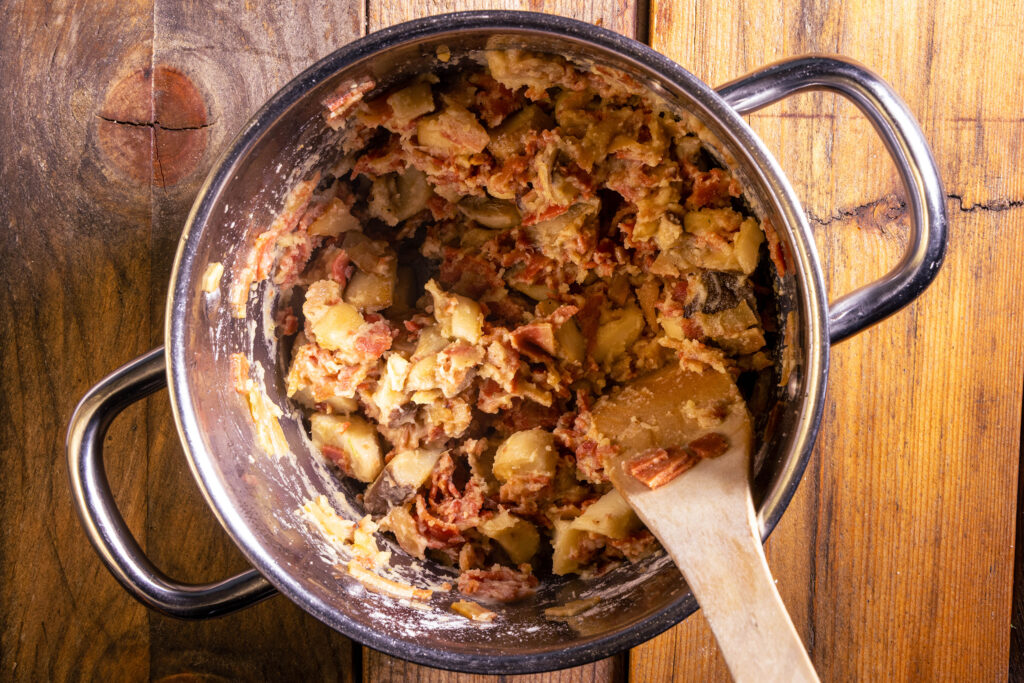 Kräuterseitlinge und veganer Schinken braten mit Mehl in einem Topf.