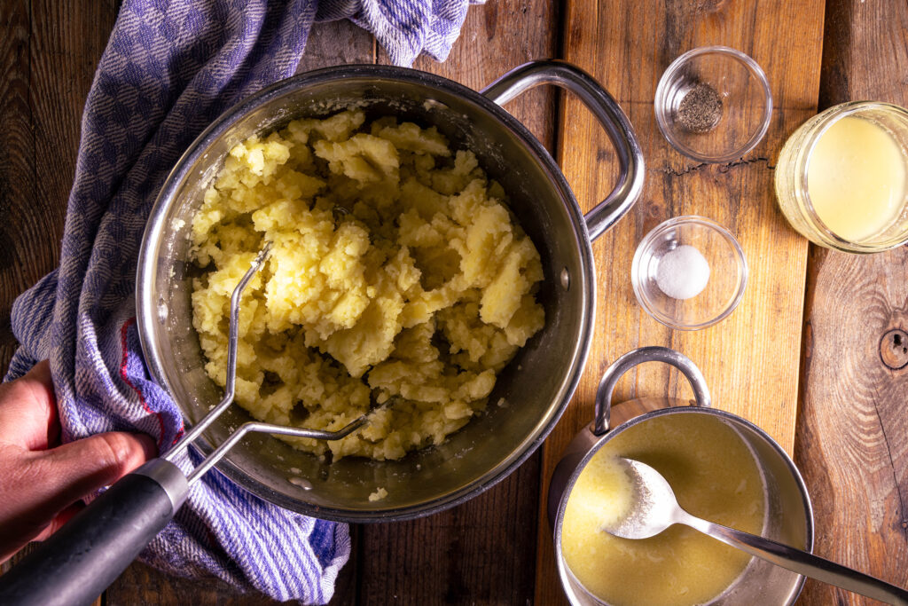 Gestampfte Kartoffeln in einem Topf, ein kleiner Topf mit geschmolzener, veganer Butter, Sojamilch und Sahne. Sowie kleine Schalen.