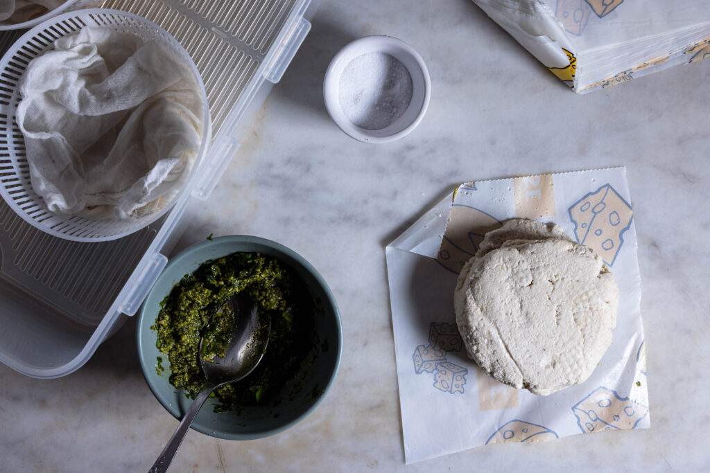 Roher veganer Cammebert auf Käsepapier, im Abseihtuch und eine Schale mit Bärlauchpesto.