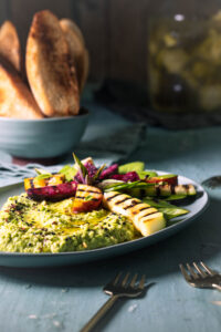 Grüner Umami-Dip aus Erbse und fermentiertem Brokkoli mit Grillgemüse auf einem Teller. Dahinter geröstetes Brot und ein Glas fermentierter Brokkoli.